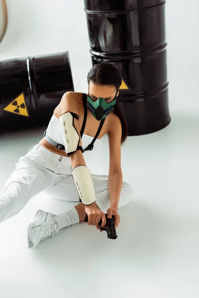 Futurista afroamericana mujer en máscara de seguridad recarga pistola cerca de los barriles de residuos radiactivos sobre fondo blanco - foto de stock