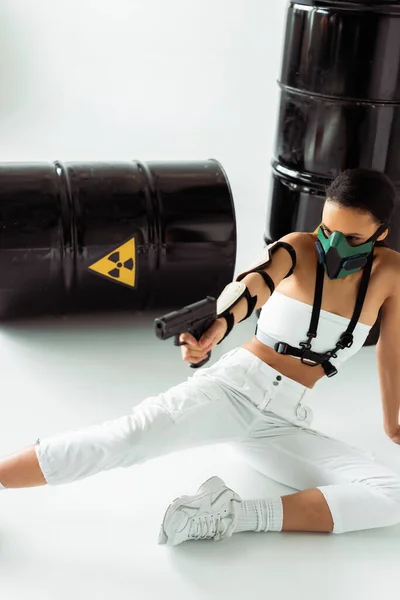 Futuristic african american woman in safety mask aiming gun near radioactive waste barrels on white background — Stock Photo