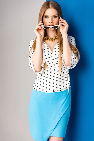 Atractiva chica posando en blusa de lunares, falda azul y gafas de sol en gris y azul - foto de stock