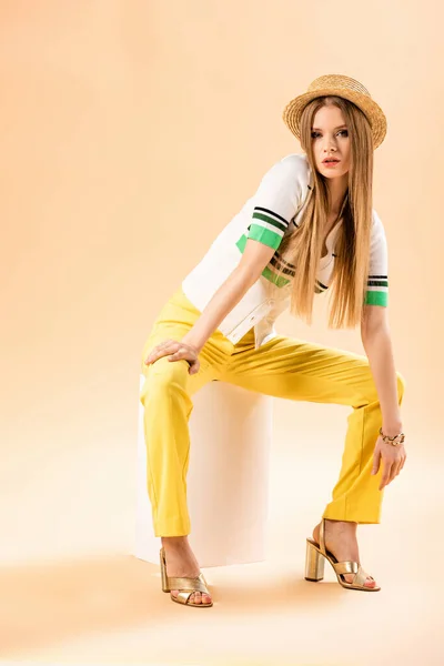 Giovane donna alla moda in pantaloni gialli, polo e cappello di paglia seduta su cubo bianco su beige — Foto stock