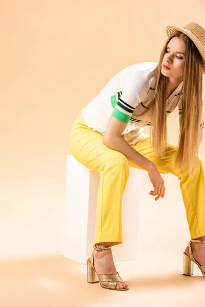 Chica con estilo en pantalones amarillos, polo y sombrero de paja sentado en cubo blanco en beige - foto de stock