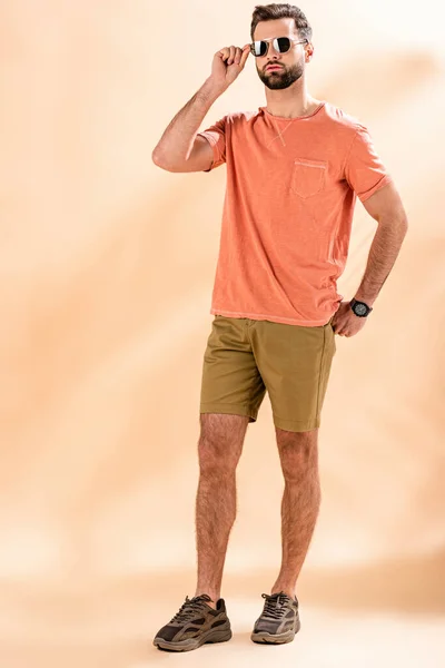 Стильний молодий чоловік позує в шортах, літня футболка і сонцезахисні окуляри на бежевому — стокове фото