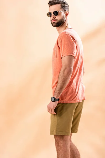 Schöner junger Mann posiert in kurzen Hosen, Sommer-T-Shirt und Sonnenbrille auf beige — Stockfoto