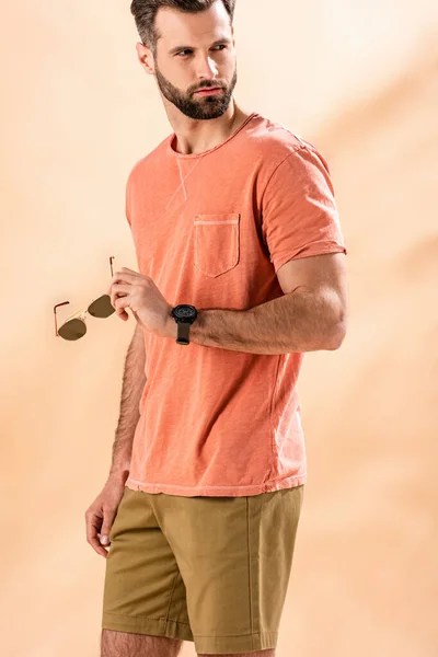 Guapo elegante hombre posando en pantalones cortos y verano camiseta sosteniendo gafas de sol en beige - foto de stock