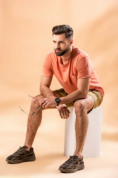 Young bearded man in shorts and summer t-shirt holding sunglasses sitting on white cube on beige — Stock Photo