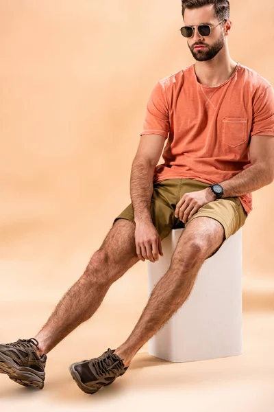 Hombre joven en pantalones cortos, camiseta de verano y gafas de sol sentado en cubo blanco en beige - foto de stock
