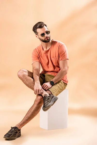 Homme élégant en short, t-shirt d'été et lunettes de soleil assis sur cube blanc sur beige — Photo de stock