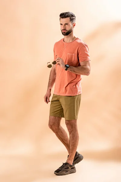 Stylish handsome man posing in shorts and summer t-shirt holding sunglasses on beige — Stock Photo