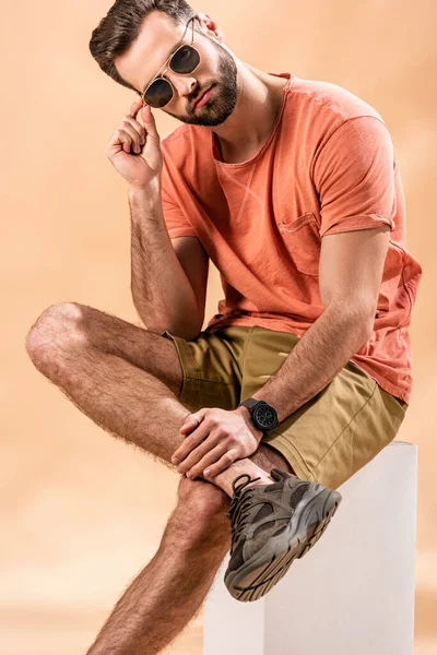 Jeune homme en short et t-shirt d'été portant des lunettes de soleil assis sur cube blanc sur beige — Photo de stock