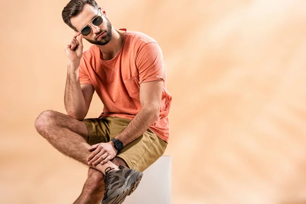 Joven guapo de moda en pantalones cortos, camiseta de verano y gafas de sol sentado en cubo blanco en beige - foto de stock