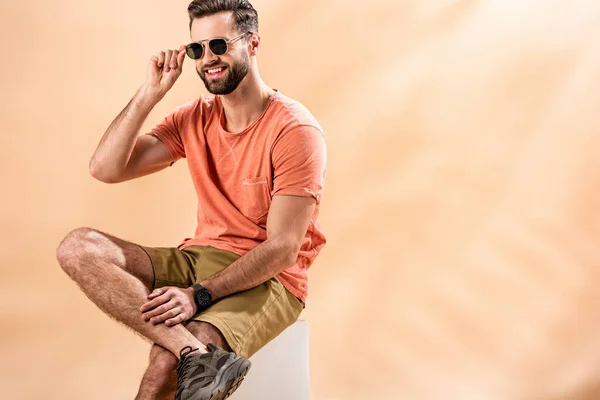 Cheerful young man in shorts, summer t-shirt and sunglasses sitting on white cube on beige — Stock Photo