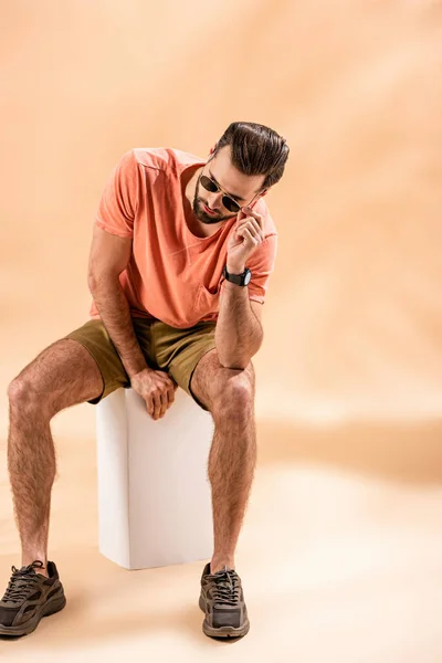 Hombre joven de moda en pantalones cortos, camiseta de verano y gafas de sol sentado en cubo blanco en beige - foto de stock