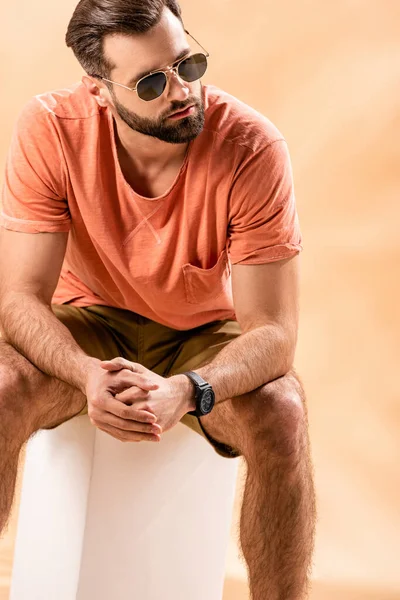 Handsome man in shorts, summer t-shirt and sunglasses sitting on white cube on beige — Stock Photo