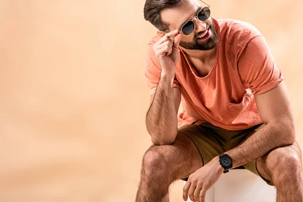 Homem bonito feliz em shorts, verão t-shirt e óculos de sol sentado no cubo branco no bege — Fotografia de Stock