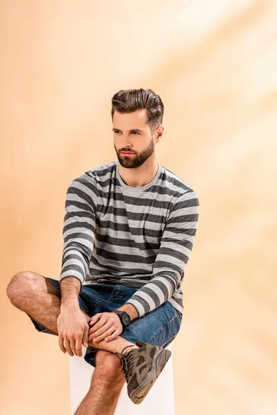 Bearded fashionable man in striped sweatshirt sitting on white cube on beige — Stock Photo