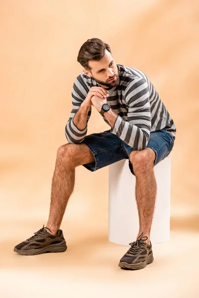 Стильный молодой человек в полосатой толстовке сидит на белом кубе на бежевом — стоковое фото