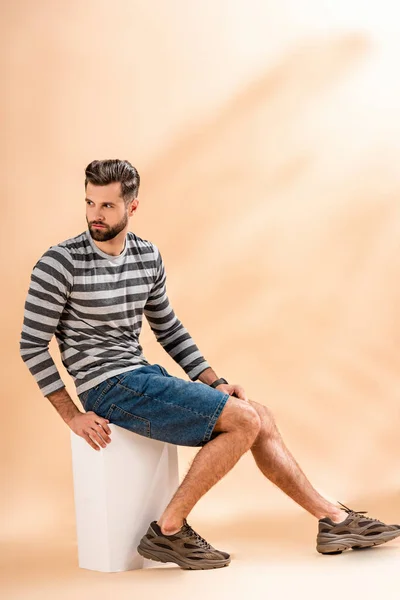 Handsome bearded man in striped sweatshirt sitting on white cube on beige — Stock Photo