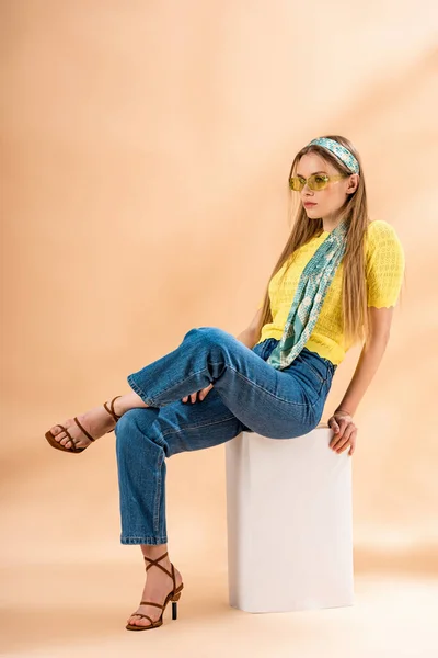 Chica rubia de moda en jeans, camiseta amarilla, gafas de sol, sandalias de tacón y bufanda de seda sentado en cubo blanco en beige - foto de stock