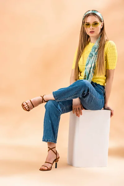 Chica rubia con estilo en jeans, camiseta amarilla, gafas de sol, sandalias de tacón y bufanda de seda sentado en cubo blanco en beige - foto de stock