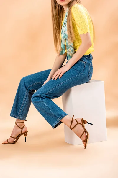 Vista recortada de niña sentada en jeans, camiseta amarilla, sandalias de tacón y bufanda de seda en beige - foto de stock