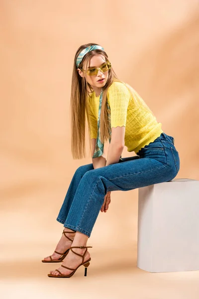 Élégante fille blonde en jeans, t-shirt jaune, lunettes de soleil, sandales à talons et foulard en soie assis sur cube blanc sur beige — Photo de stock