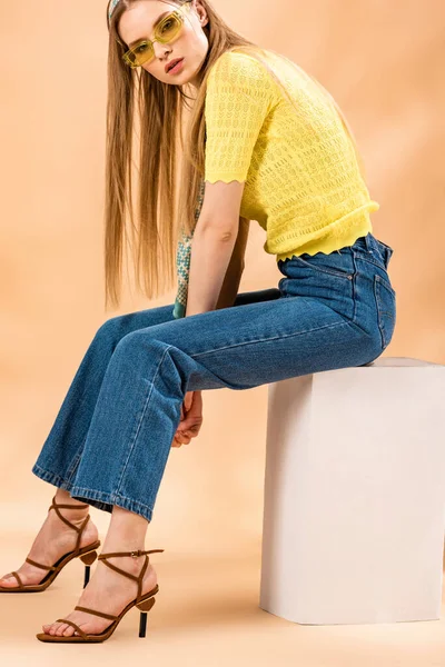 Модная блондинка в джинсах, желтая футболка, солнечные очки, сандалии на каблуках и шелковый шарф, сидящий на белом кубе на бежевом — стоковое фото