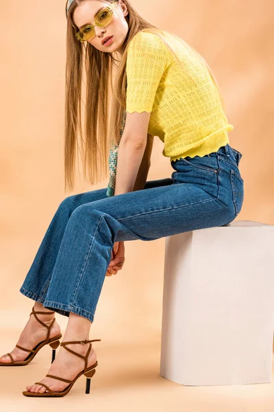 Menina loira na moda em jeans, camiseta amarela, óculos de sol, sandálias de salto alto e cachecol de seda sentado no cubo branco no bege — Fotografia de Stock