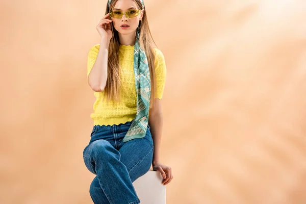 Chica de moda en jeans, camiseta amarilla, gafas de sol y bufanda de seda sentado en cubo blanco en beige - foto de stock