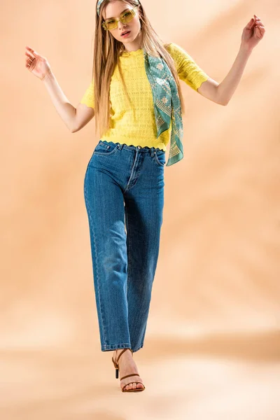 Приваблива дівчина позує в джинсах, жовтій футболці, сонцезахисних окулярах і шовковому шарфі на бежевому — стокове фото