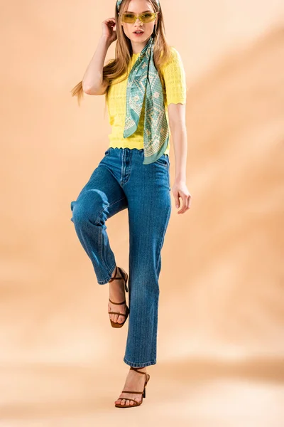 Attractive girl posing in jeans, yellow t-shirt, sunglasses and silk scarf on beige — Stock Photo