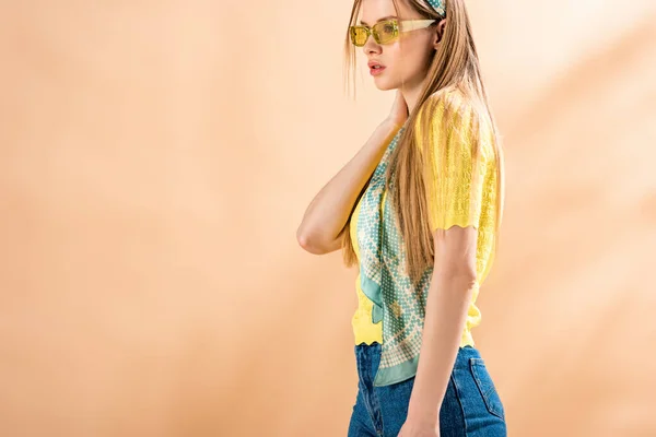 Beautiful fashionable girl posing in jeans, yellow t-shirt, sunglasses and silk scarf on beige — Stock Photo