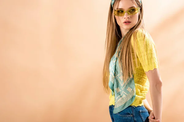 Красива стильна дівчина позує в джинсах, жовтій футболці, сонцезахисних окулярах і шовковому шарфі на бежевому — стокове фото
