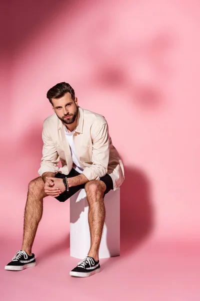 Fashionable man in summer shirt and shorts sitting on white cube on pink — Stock Photo