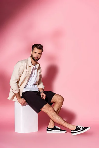 Guapo hombre con estilo en camisa de verano y pantalones cortos sentados en cubo blanco en rosa - foto de stock