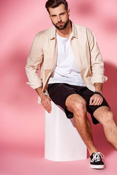 Uomo barbuto alla moda in posa in camicia estiva e pantaloncini su cubo bianco su rosa — Foto stock