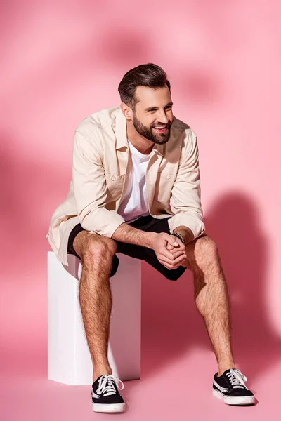 Uomo barbuto sorridente in camicia estiva e pantaloncini seduti su cubo bianco su rosa — Foto stock