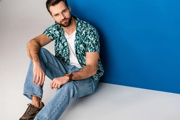 Stylish young man posing in summer clothes and sunglasses on grey and blue — Stock Photo