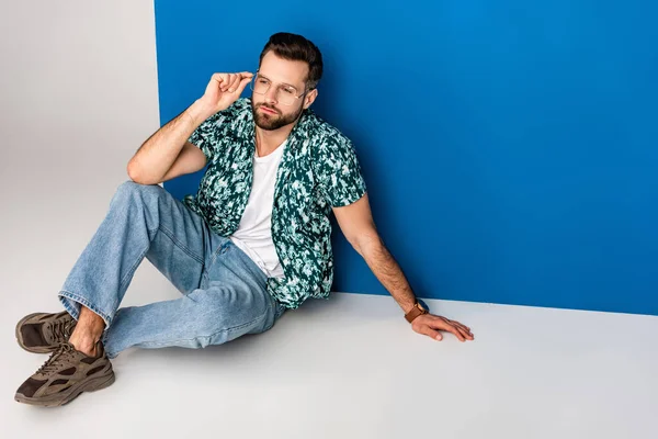 Bearded young man posing in summer clothes and sunglasses on grey and blue — Stock Photo