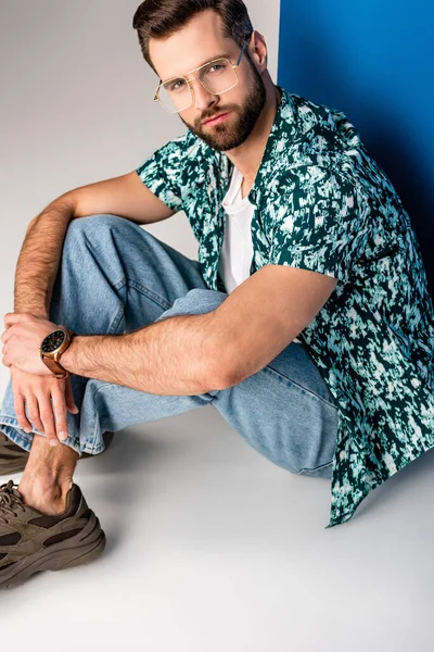 Stylischer junger Mann posiert in Sommerkleidung und Sonnenbrille auf grau und blau — Stockfoto