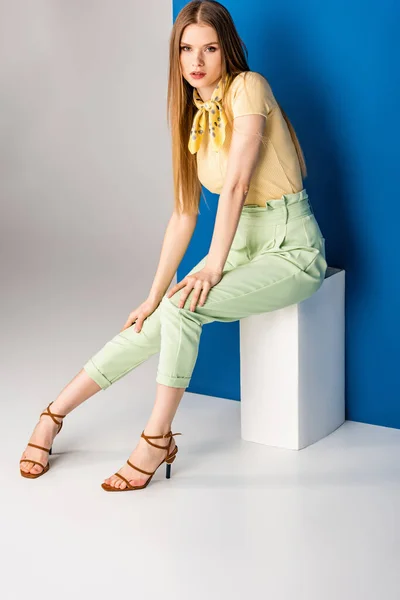 Hermosa mujer en pantalones verdes de verano y sandalias de tacón sentado en cubo blanco en gris y azul - foto de stock