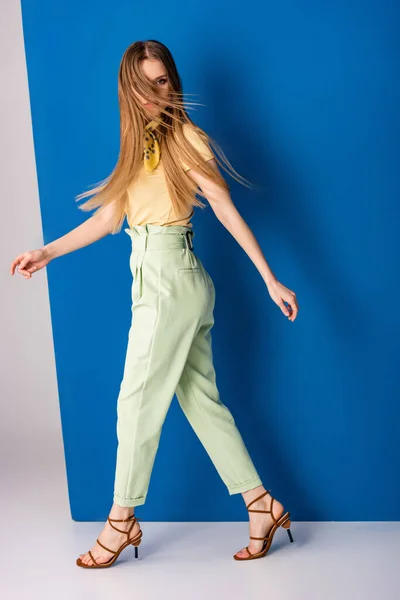 Красивая модная девушка позирует в зеленых летних брюках и сандалиях на каблуках на сером и голубом — стоковое фото