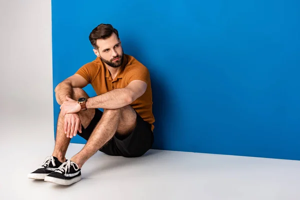 Stylish handsome man sitting in shorts and brown polo on grey and blue — Stock Photo