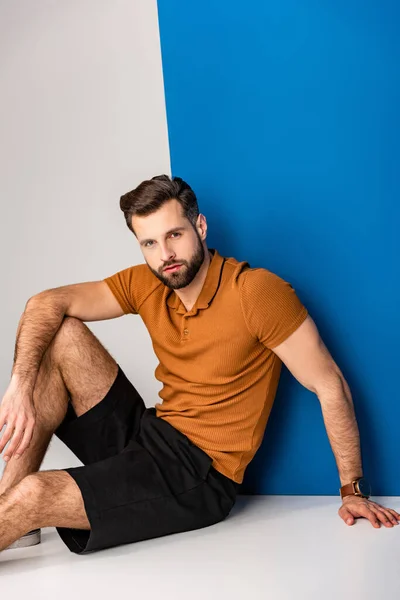 Joven de moda posando en pantalones cortos y polo marrón en gris y azul - foto de stock