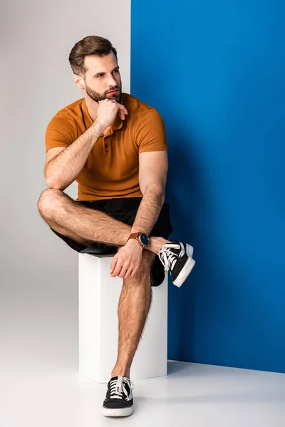 Pensive bearded man in shorts and brown polo sitting on white cube on grey and blue — Stock Photo