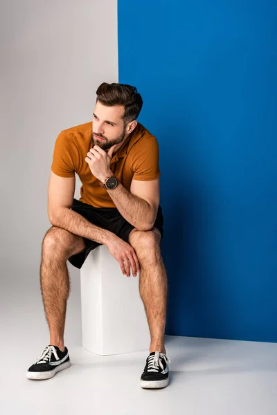 Thoughtful stylish bearded man in shorts and brown polo sitting on white cube on grey and blue — Stock Photo