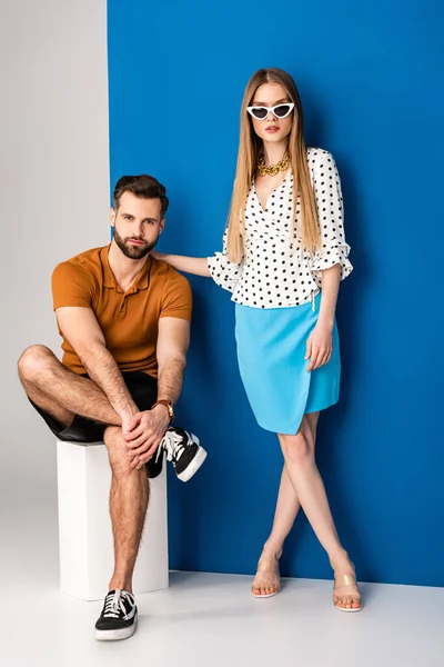 Pareja joven de moda en ropa de verano y gafas de sol posando cerca del cubo blanco en gris y azul - foto de stock