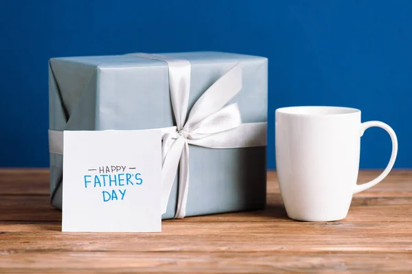 Foyer sélectif de carte de vœux avec lettrage heureux jour des pères, tasse blanche et boîte cadeau avec arc blanc isolé sur bleu — Photo de stock