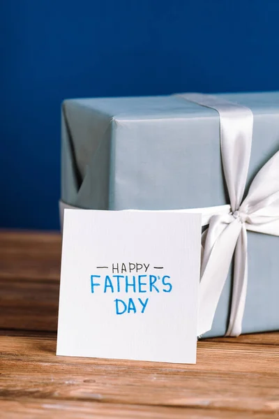 Вибірковий фокус вітальної листівки з написом щасливих батьків день і подарункова коробка з білим бантом на дерев'яному столі ізольовані на синьому — стокове фото
