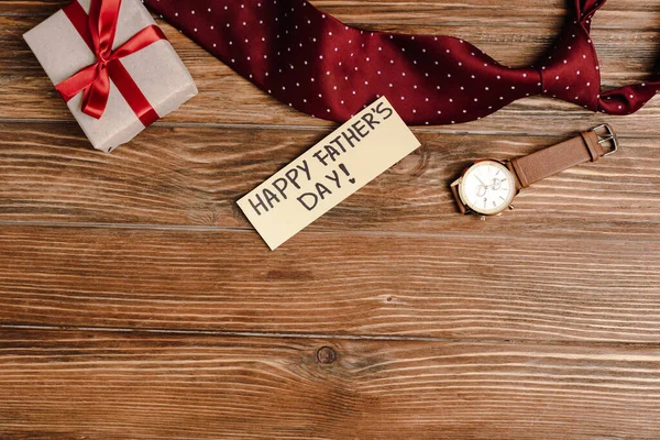Vista superior de la caja de regalo con lazo rojo, tarjeta de felicitación con letras feliz día de los padres y mens corbata y reloj de pulsera sobre fondo de madera - foto de stock