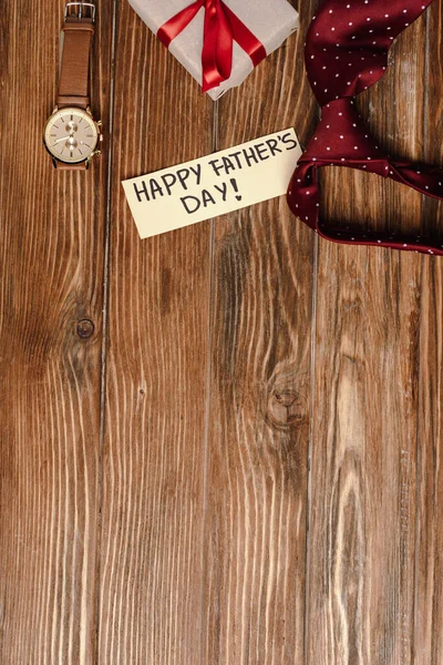 Vista dall'alto della confezione regalo con fiocco rosso, biglietto di auguri con lettering happy father day e cravatta rossa da uomo e orologio da polso su sfondo in legno — Foto stock